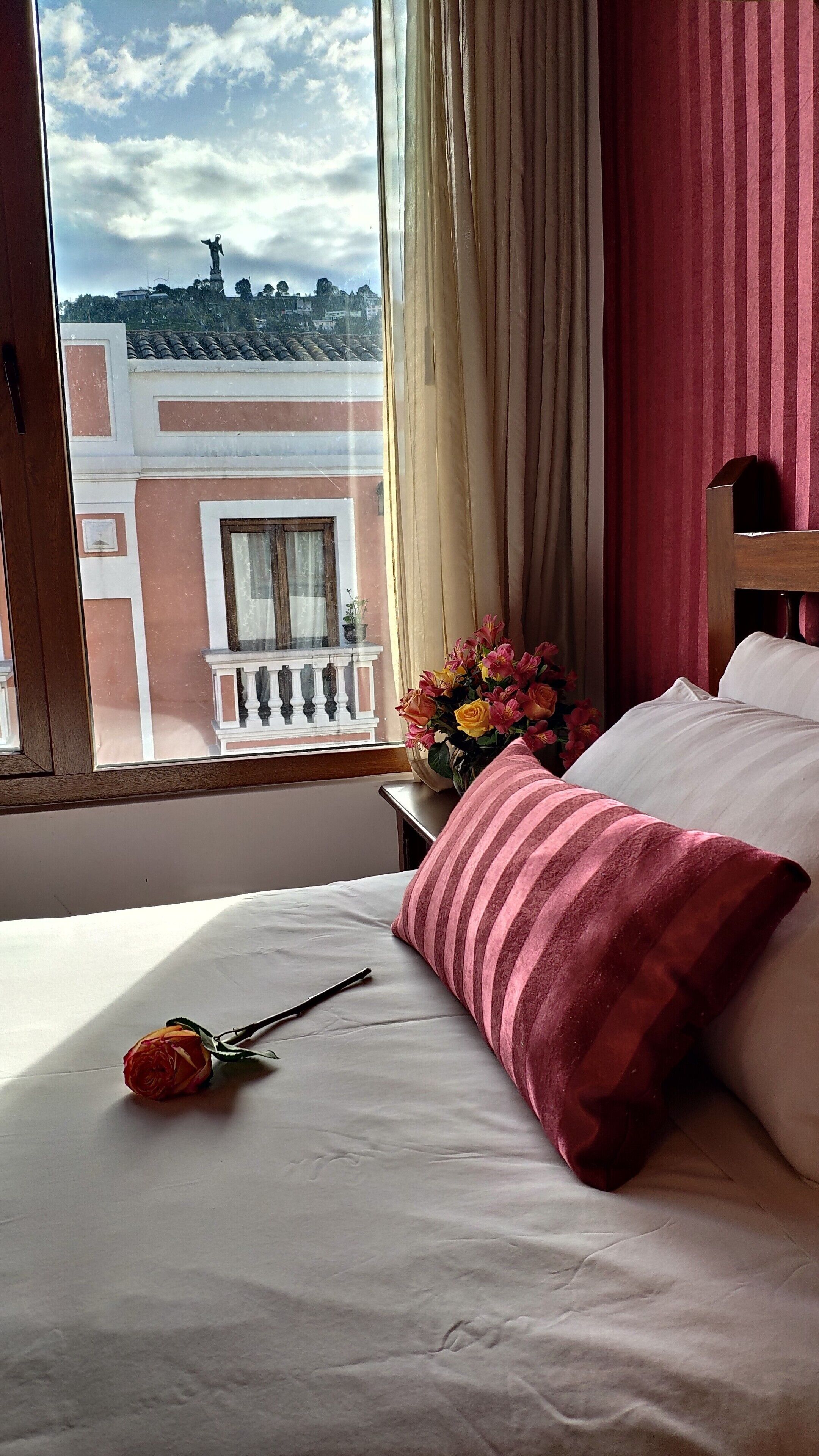 Een hotelkamer met een bed, een raam met gordijnen, een vaas met bloemen en een balkon met een beeld.