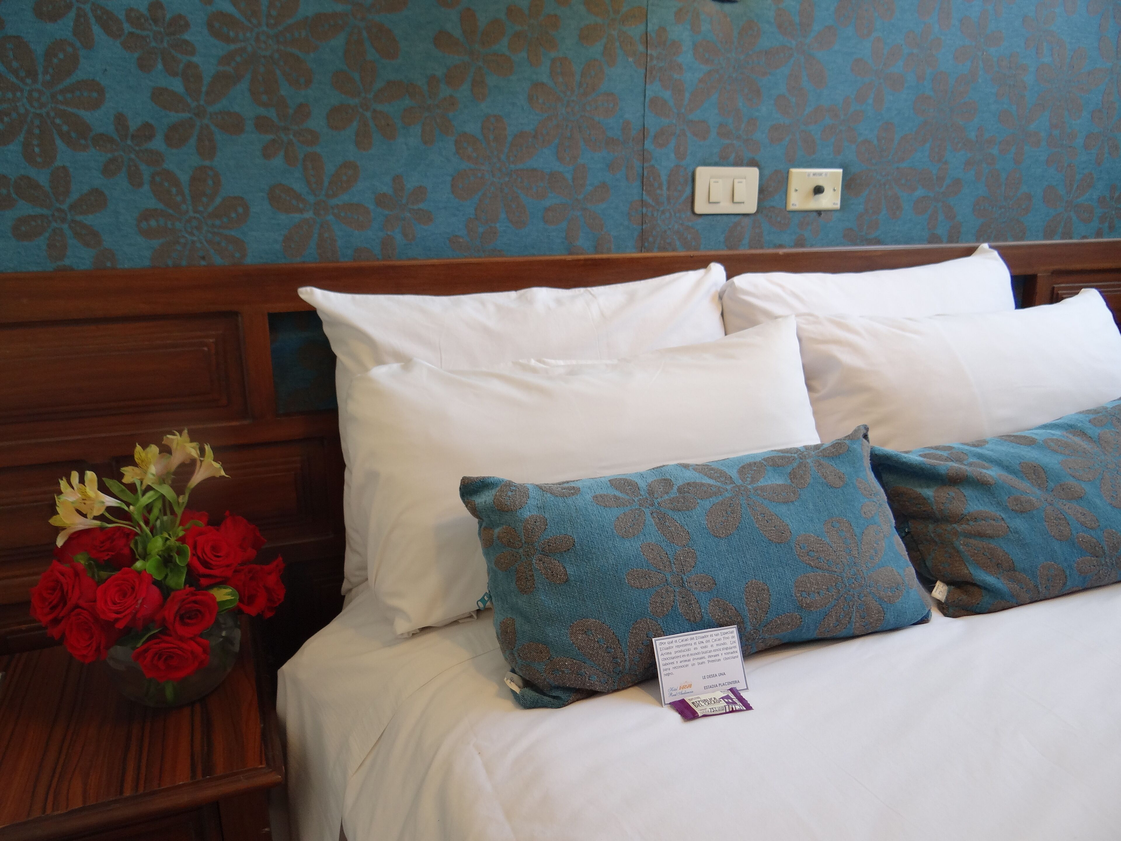 Une chambre d’hôtel avec un lit, des oreillers blancs, un coussin à motifs floraux bleus et un vase contenant des fleurs rouges et jaunes.