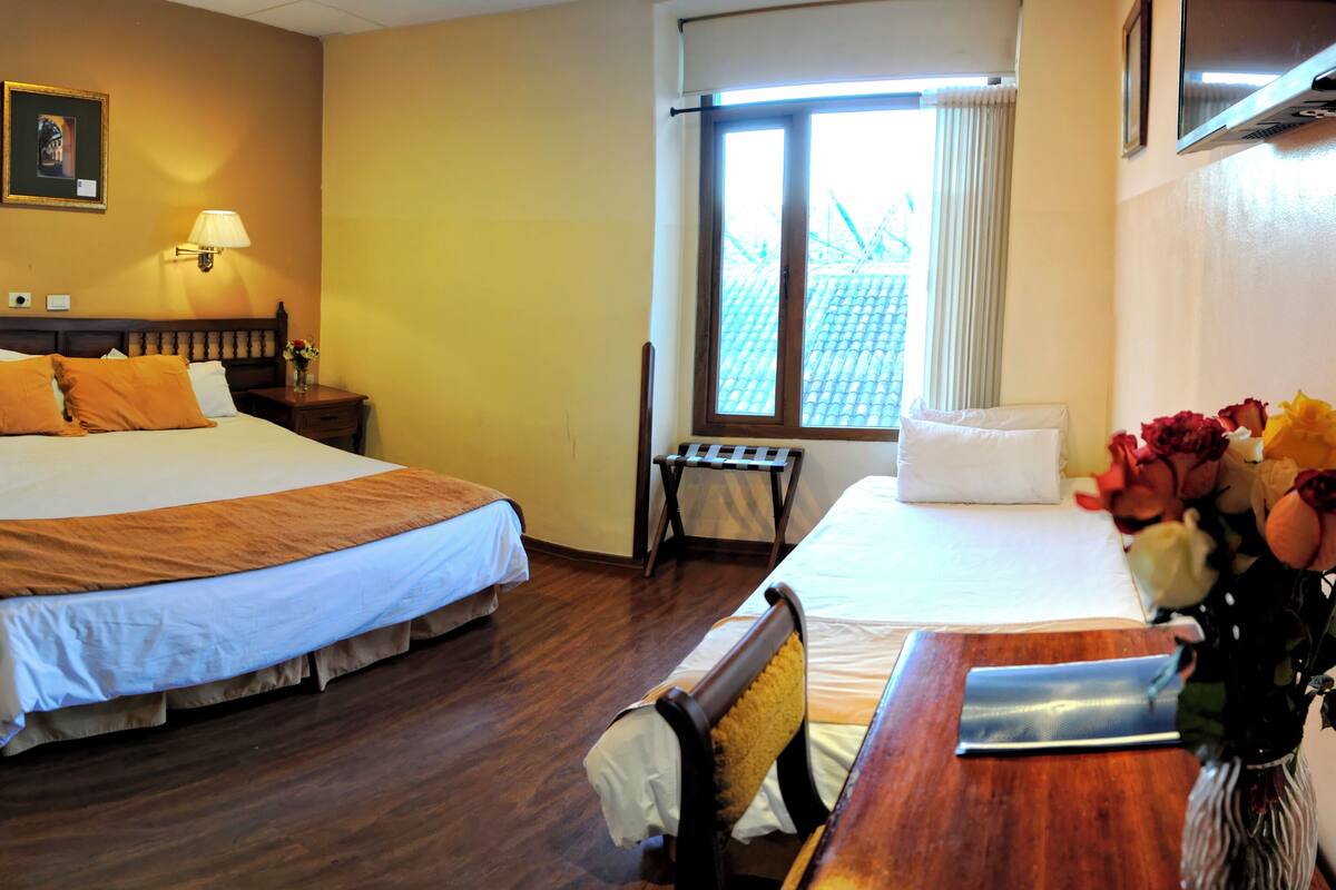 A hotel room with two beds, a wooden headboard, a nightstand, a desk, a chair, a window with curtains, and a vase with flowers.