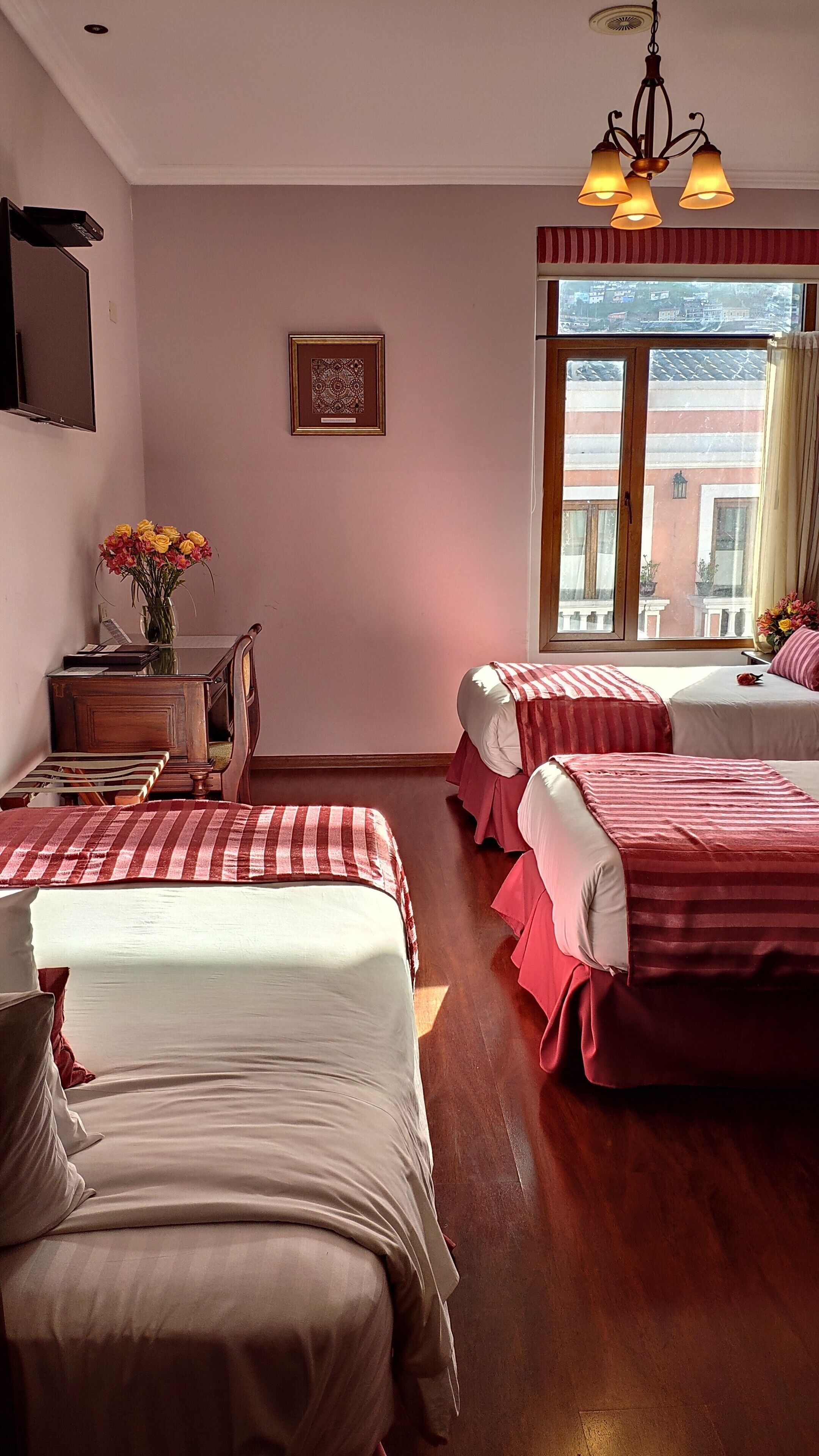 Une chambre d’hôtel avec deux lits, un bureau, une chaise, une petite table, un vase avec des fleurs et une fenêtre donnant sur des immeubles.