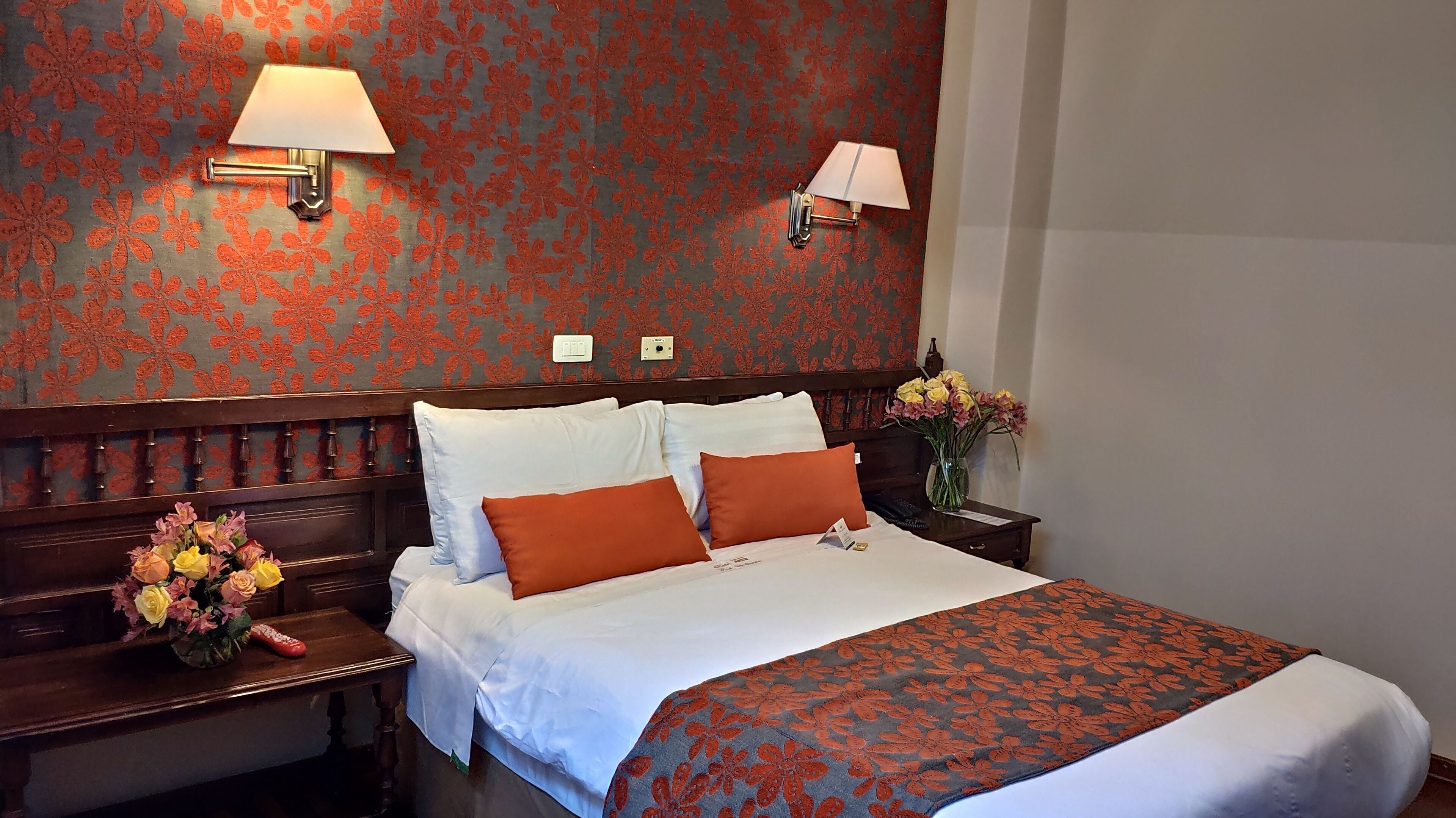 A hotel room with a large bed, two bedside tables, a nightstand, a floral-patterned wall, and two wall-mounted lamps.