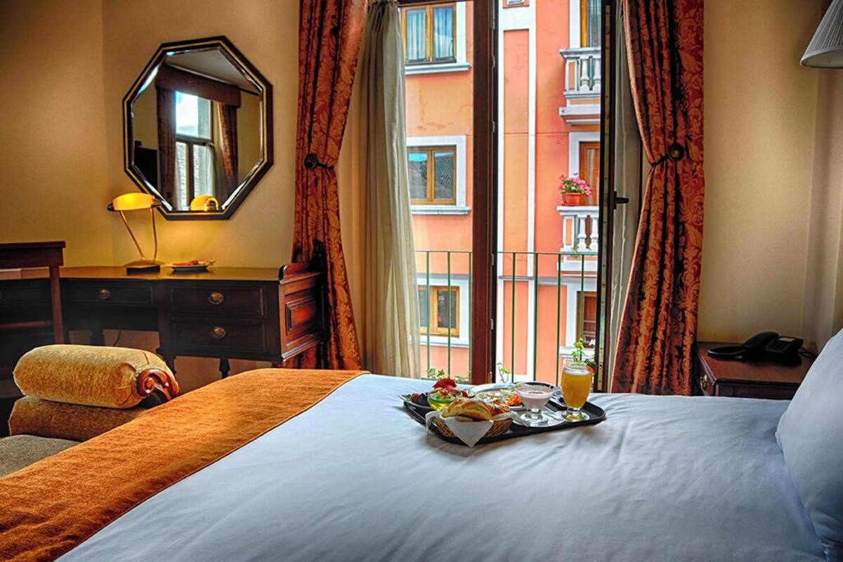 A hotel room with a bed, a bedside table with a lamp, a tray with breakfast, and a view of a building through the window.