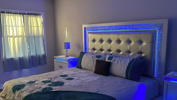 Een moderne slaapkamer met een gestoffeerd hoofdeinde, een groot bed met een bloemenprintendekbed en blauwe LED-verlichting langs de voeteneind.'
