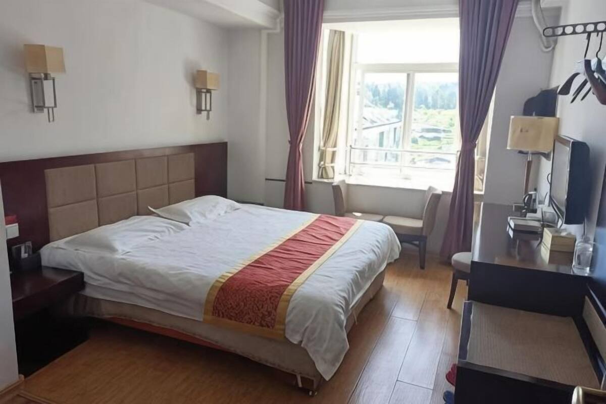 A hotel room with a large bed, a desk with a chair, a window with curtains, and a wall-mounted lamp.