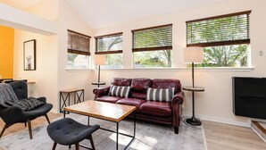 Un salon moderne avec un canapé en cuir, deux fauteuils, une table basse et un téléviseur. On y trouve trois fenêtres équipées de stores, et une lampe sur pied est visible.