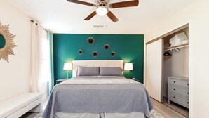 Une chambre à coucher comprenant un lit, deux lampes de chevet, un ventilateur de plafond, un élément décoratif fixé au mur et une armoire avec des vêtements suspendus.