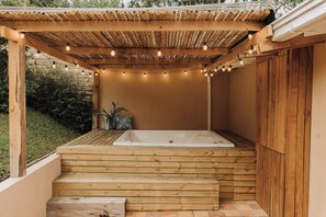 Un jacuzzi de madera bajo una pérgola con luces colgantes, rodeado de una terraza de madera y un jardín.