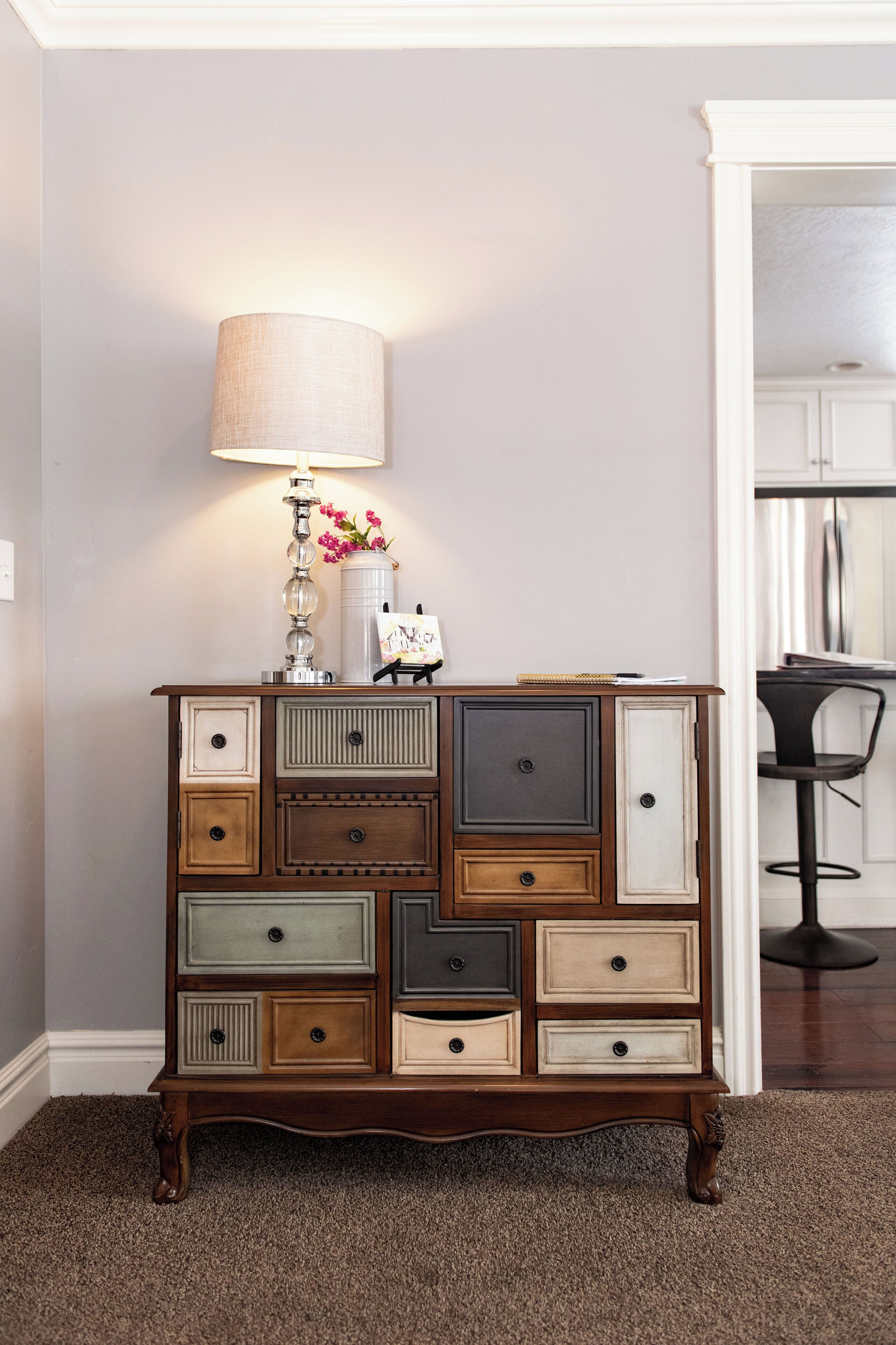 A wooden dresser with multiple drawers and a lamp on top, placed against a wall with a chair and table in the background.