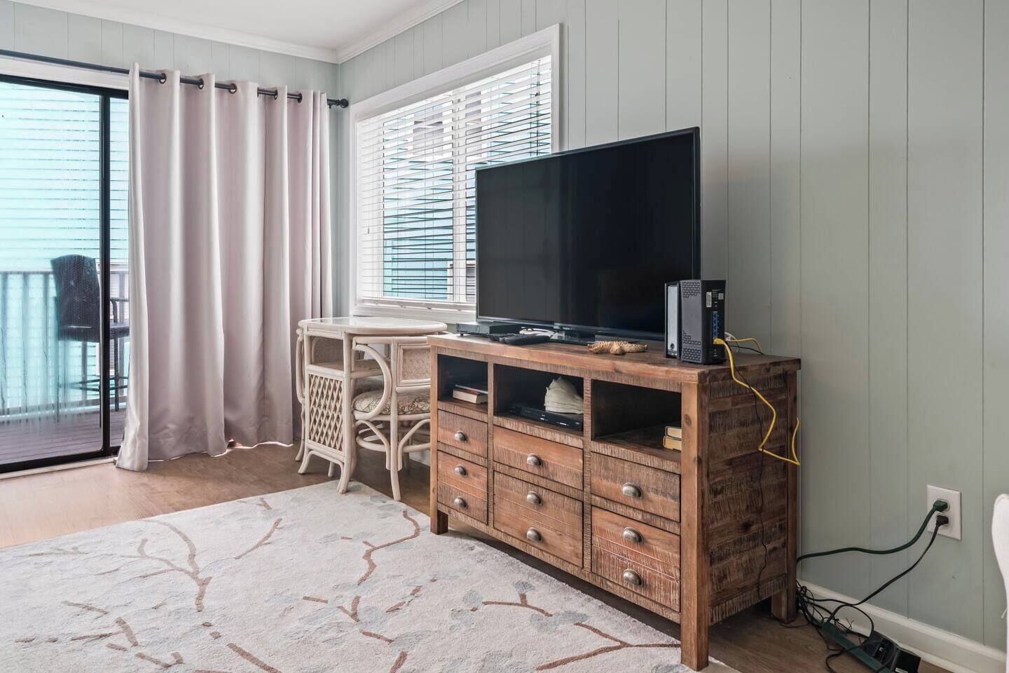 A room with a wooden TV stand, a flat-screen TV, a wicker chair, a table, and a view through a window with blinds.