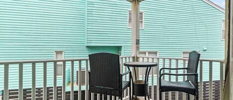 A covered balcony with two wicker chairs and a small table, overlooking a turquoise building.
