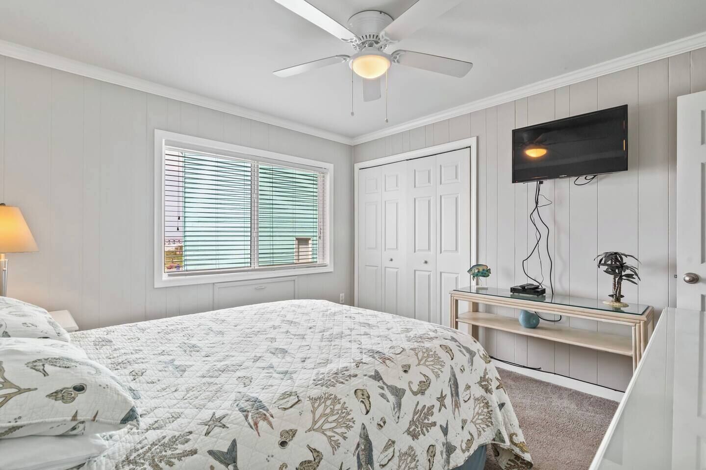 A bedroom with a bed, a ceiling fan, a TV mounted on the wall, and a window with blinds.