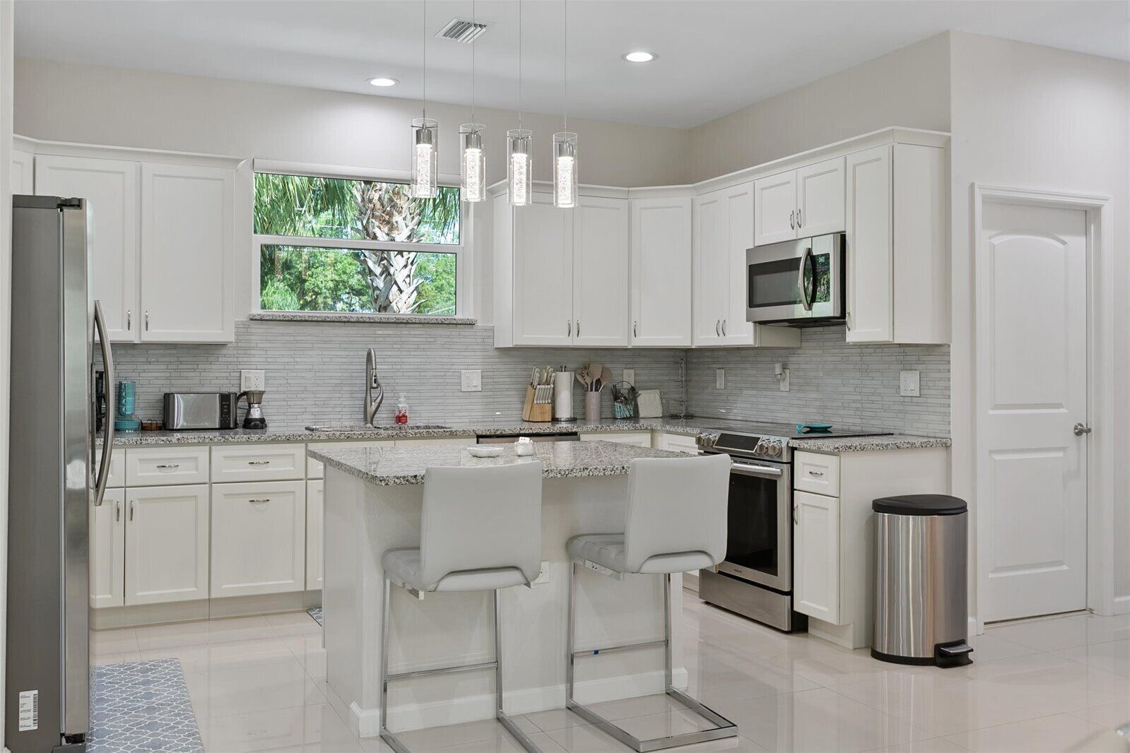 A modern kitchen with white cabinetry, a central island with bar stools, stainless steel appliances, and a window with a view of greenery.