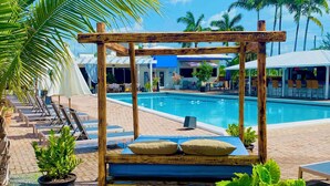Un área junto a la piscina con una cama con dosel de madera, sillones de descanso y plantas en macetas.