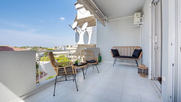 Un balcón con dos sillas de mimbre, una mesita pequeña y una silla colgante. Hay un toldo blanco que proporciona sombra, y se ve una vista del paisaje urbano al fondo.
