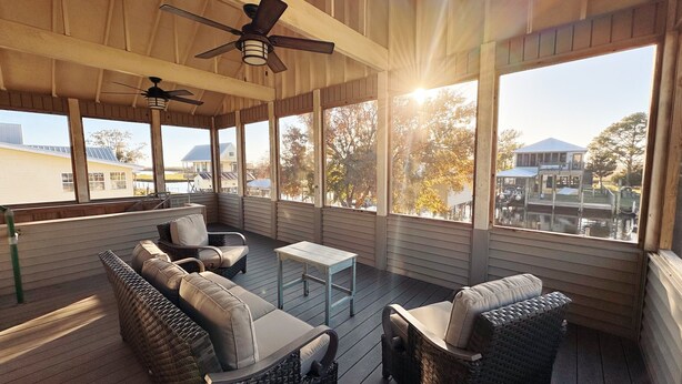 Une terrasse extérieure couverte, aménagée avec des meubles en osier, des ventilateurs de plafond et de grandes fenêtres offrant une vue sur un quartier résidentiel et sur l’eau.