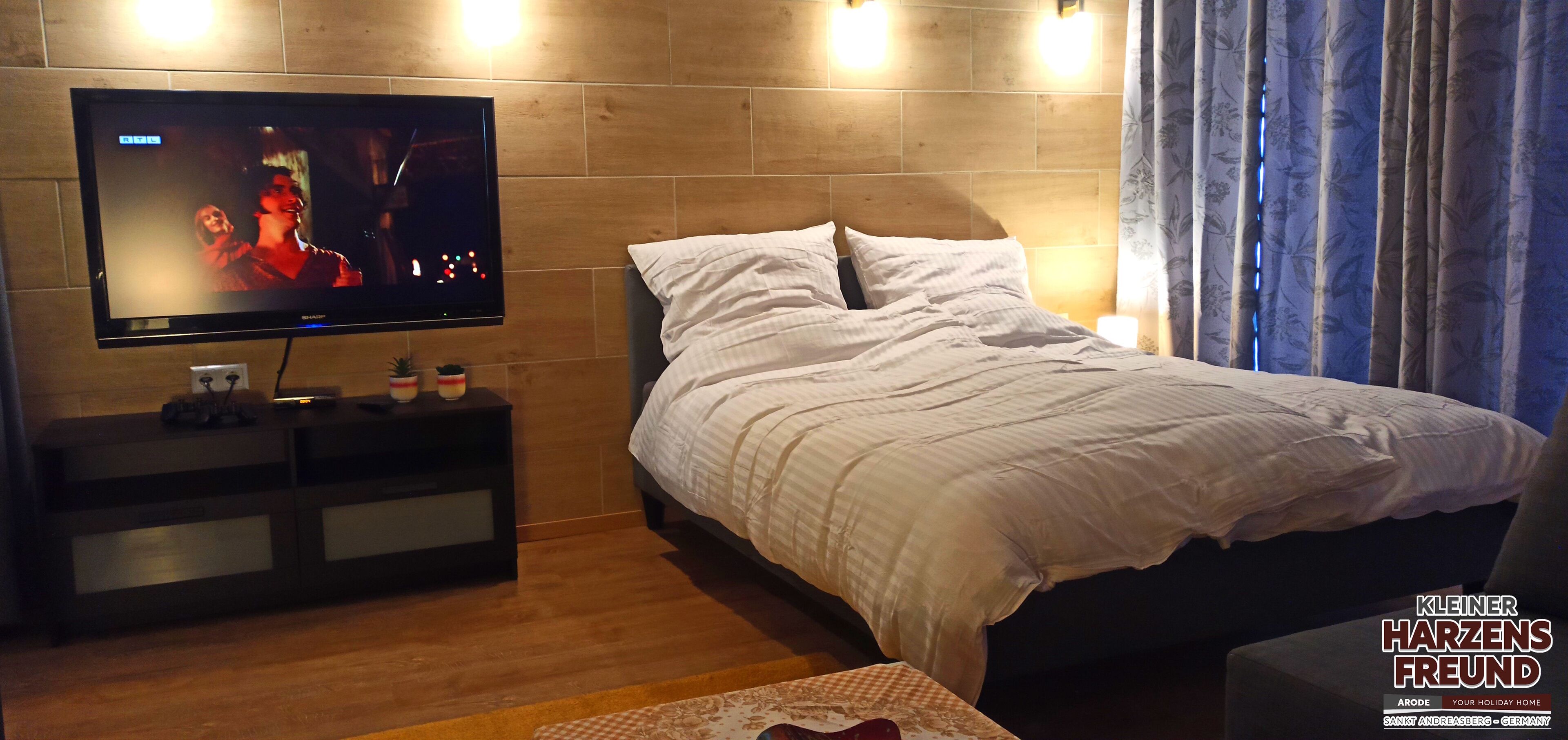 Ett hotellrum med en säng, en tv på ett stativ, ett nattduksbord och ett fönster med gardiner.
