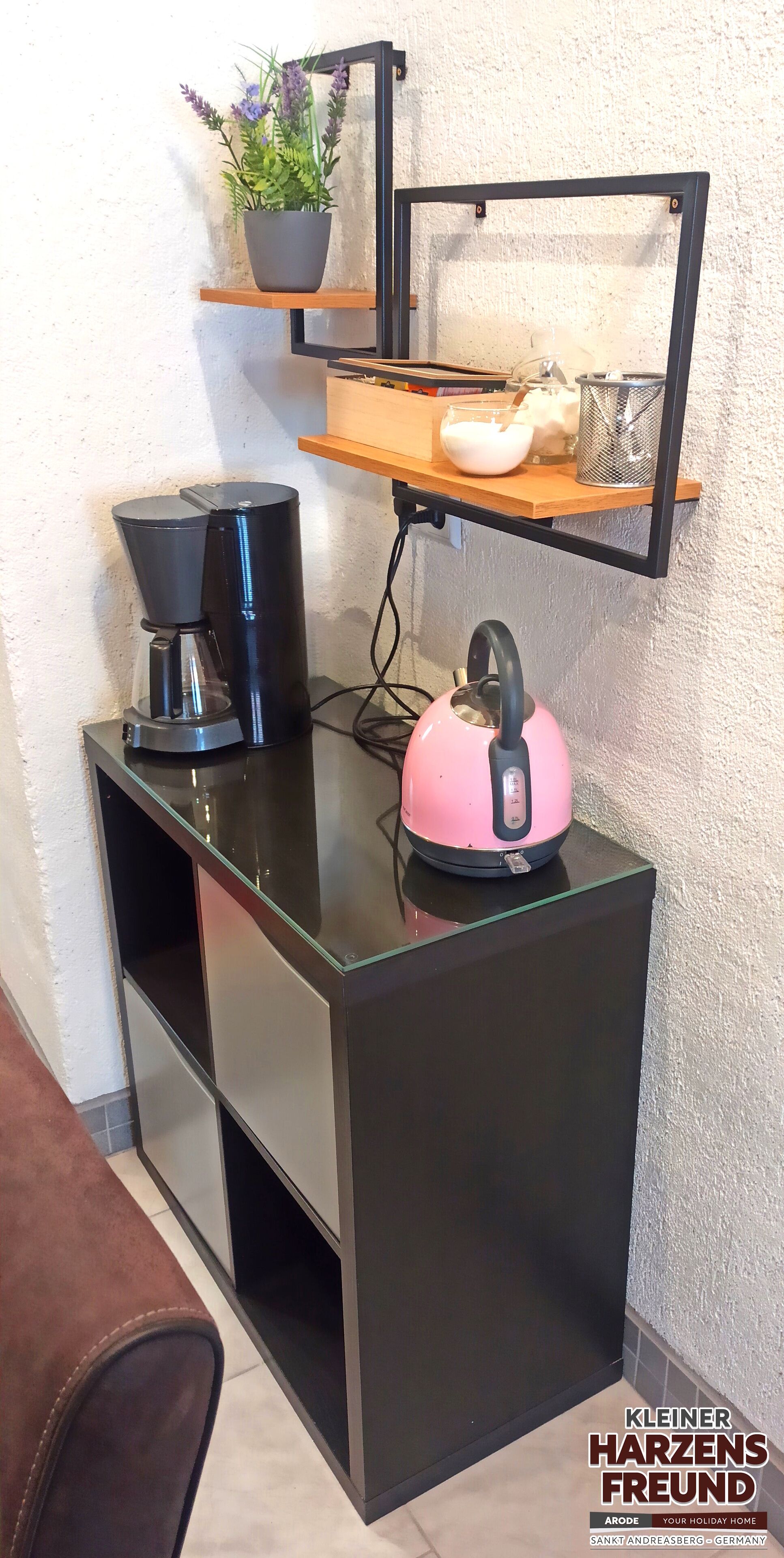 Una kitchenette compacta con cafetera, hervidor y una planta en maceta sobre una estantería.