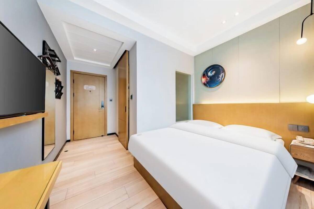 A modern hotel room with a large bed, a flat-screen TV mounted on the wall, a bedside table, and a wooden floor.