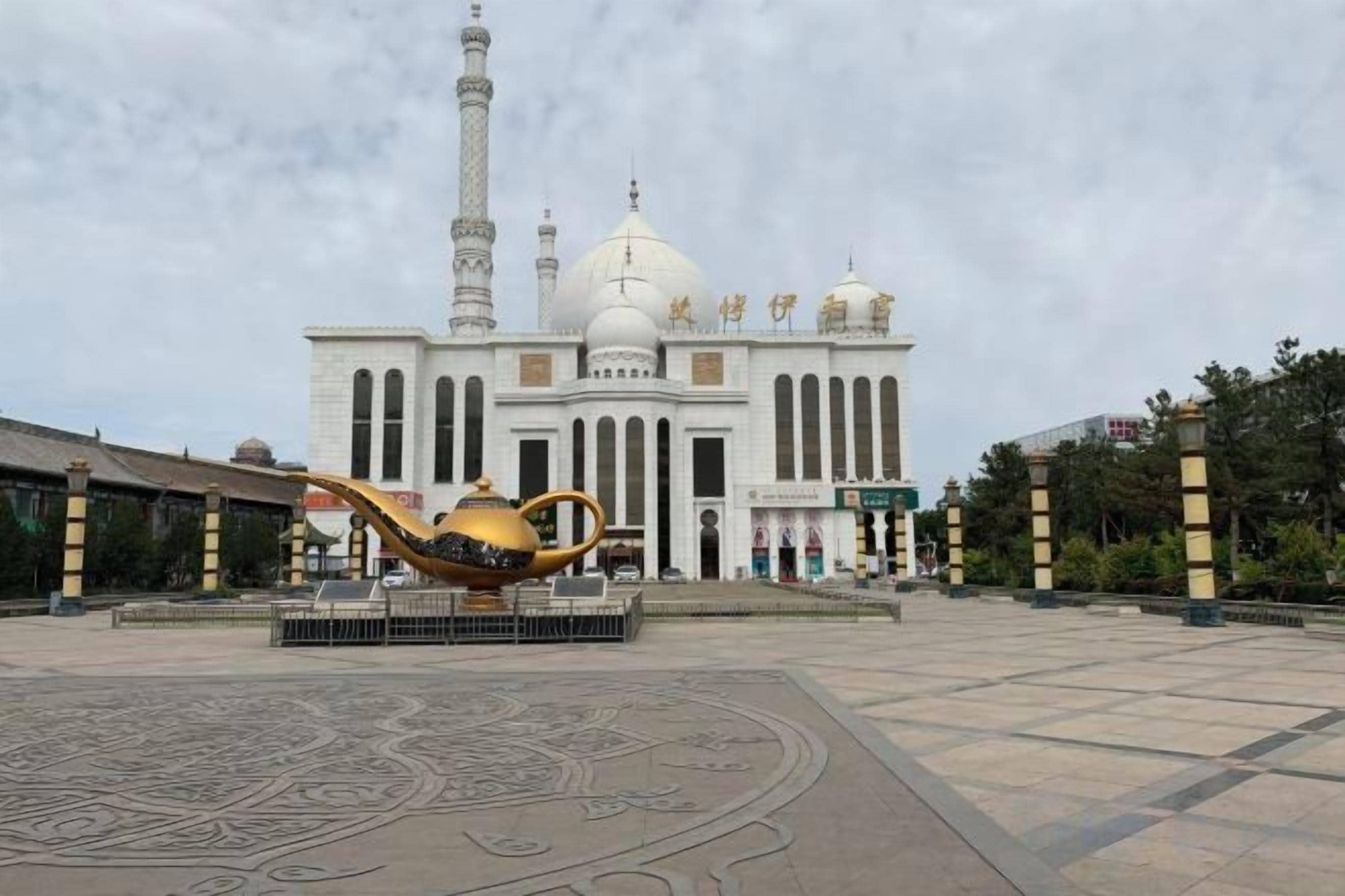 A large building with a prominent minaret and a dome, surrounded by a plaza with a decorative golden lamp.