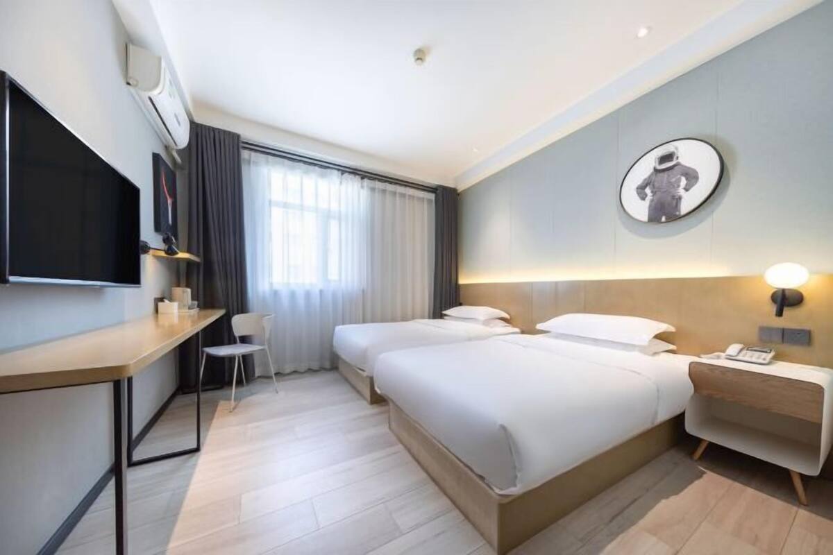 A hotel room with a bed, bedside table, lamp, TV, and a desk with a chair.