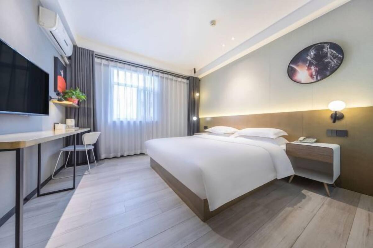 A hotel room with a bed, bedside table, chair, TV, and a large window with curtains.