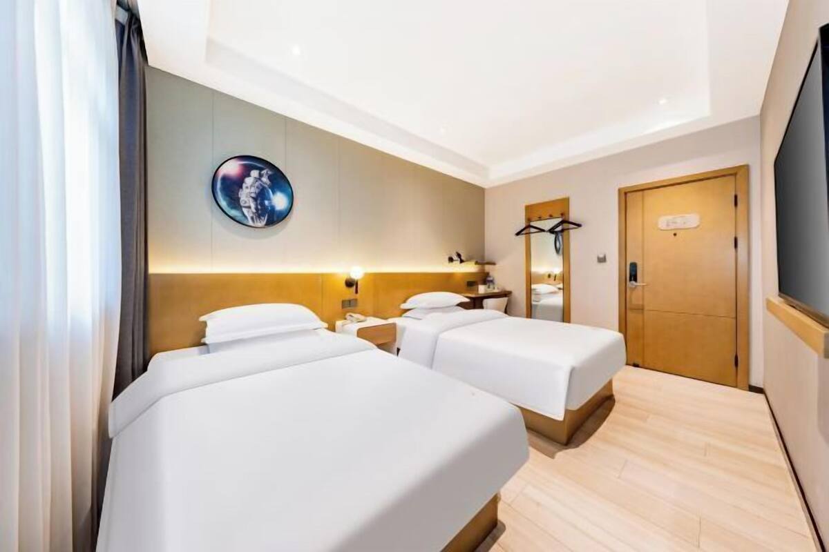 A hotel room with two beds, a bedside table, a mirror, a coat rack, and a wall-mounted TV.