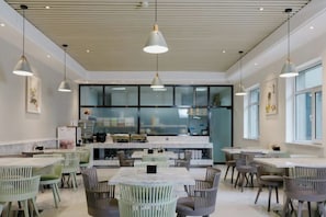 Un comedor moderno con mesas y sillas, una cocina al fondo e iluminación colgante.