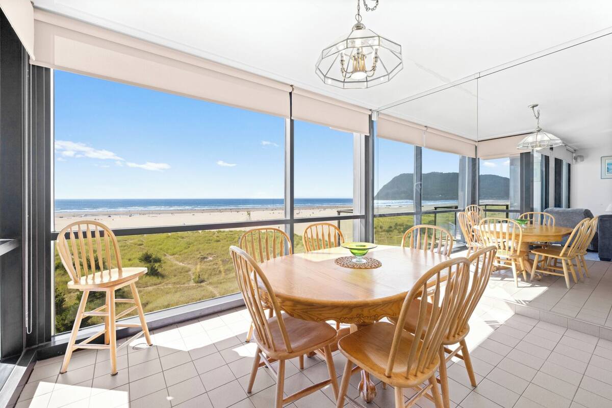 Un'ampia sala da pranzo con un tavolo grande e sedie, ampie finestre che si affacciano sulla spiaggia e sull'oceano, e un lampadario moderno.