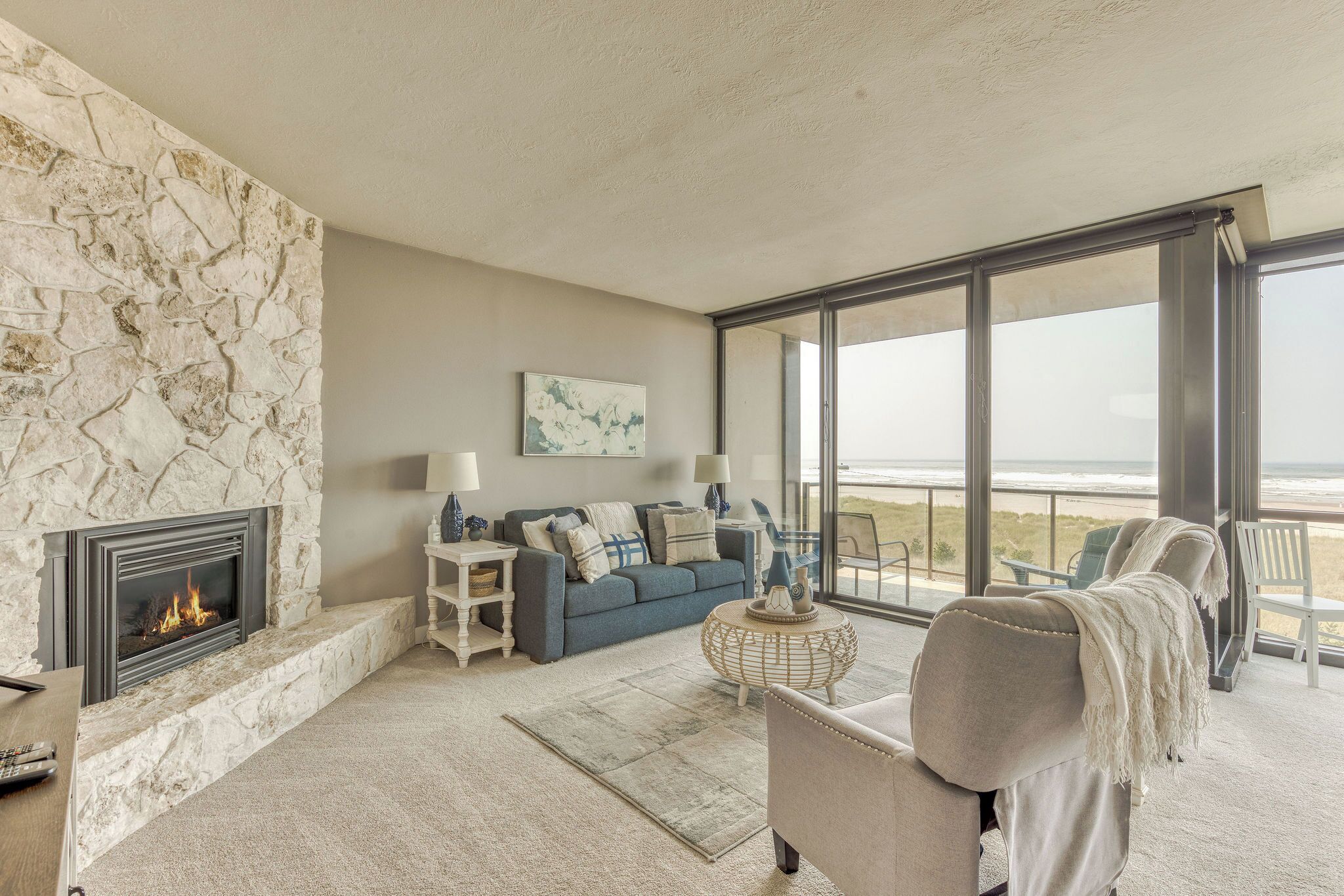 Un salón con chimenea de piedra, un sofá azul y vistas a la playa a través de amplios ventanales.
