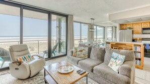 Un vaste espace de vie comprenant un canapé, un fauteuil inclinable et une table basse. Une grande fenêtre offre une vue sur la plage.