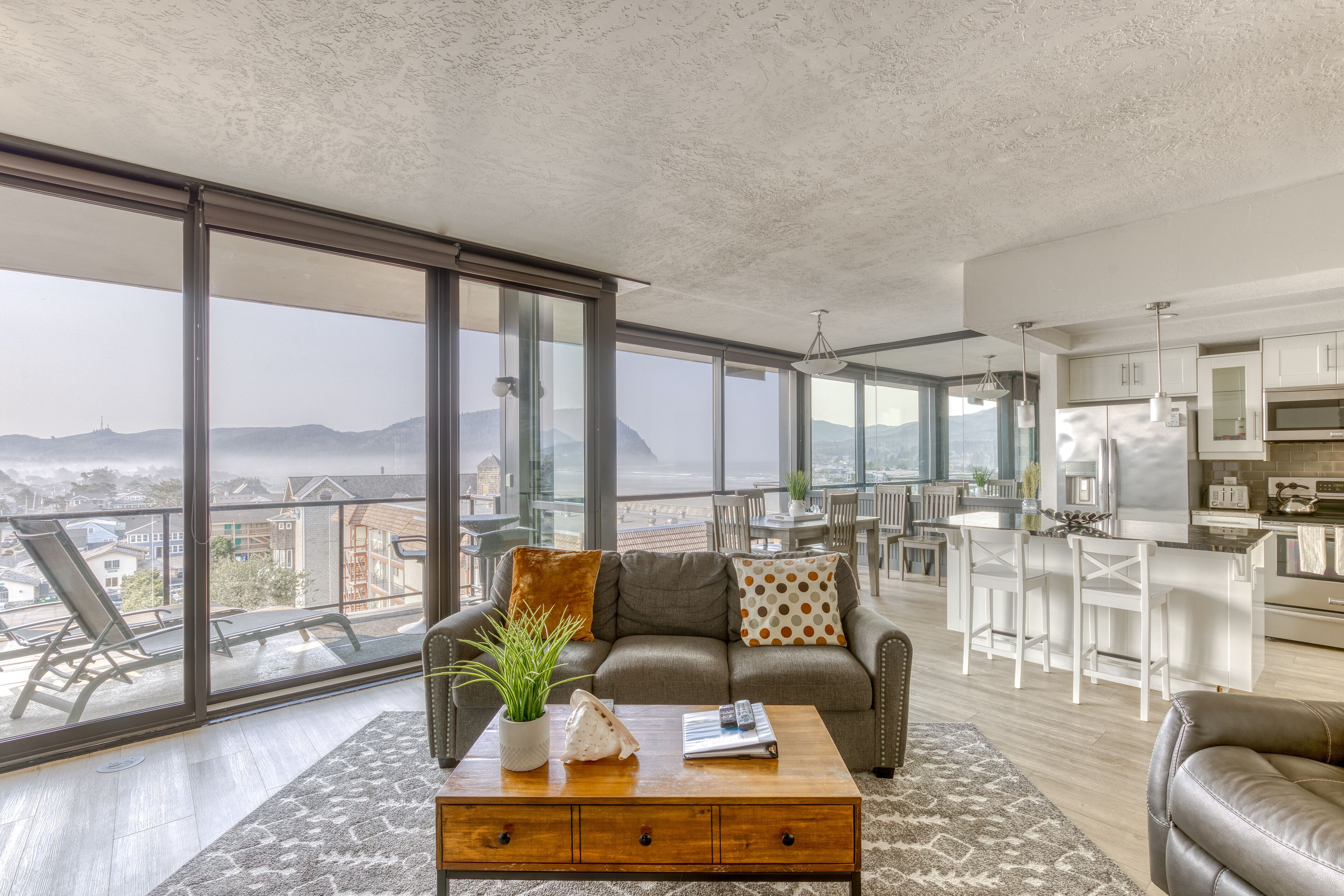 Un vaste espace de vie avec une grande fenêtre donnant sur le paysage urbain, un coin salon confortable avec une table basse et une cuisine moderne aux meubles blancs.