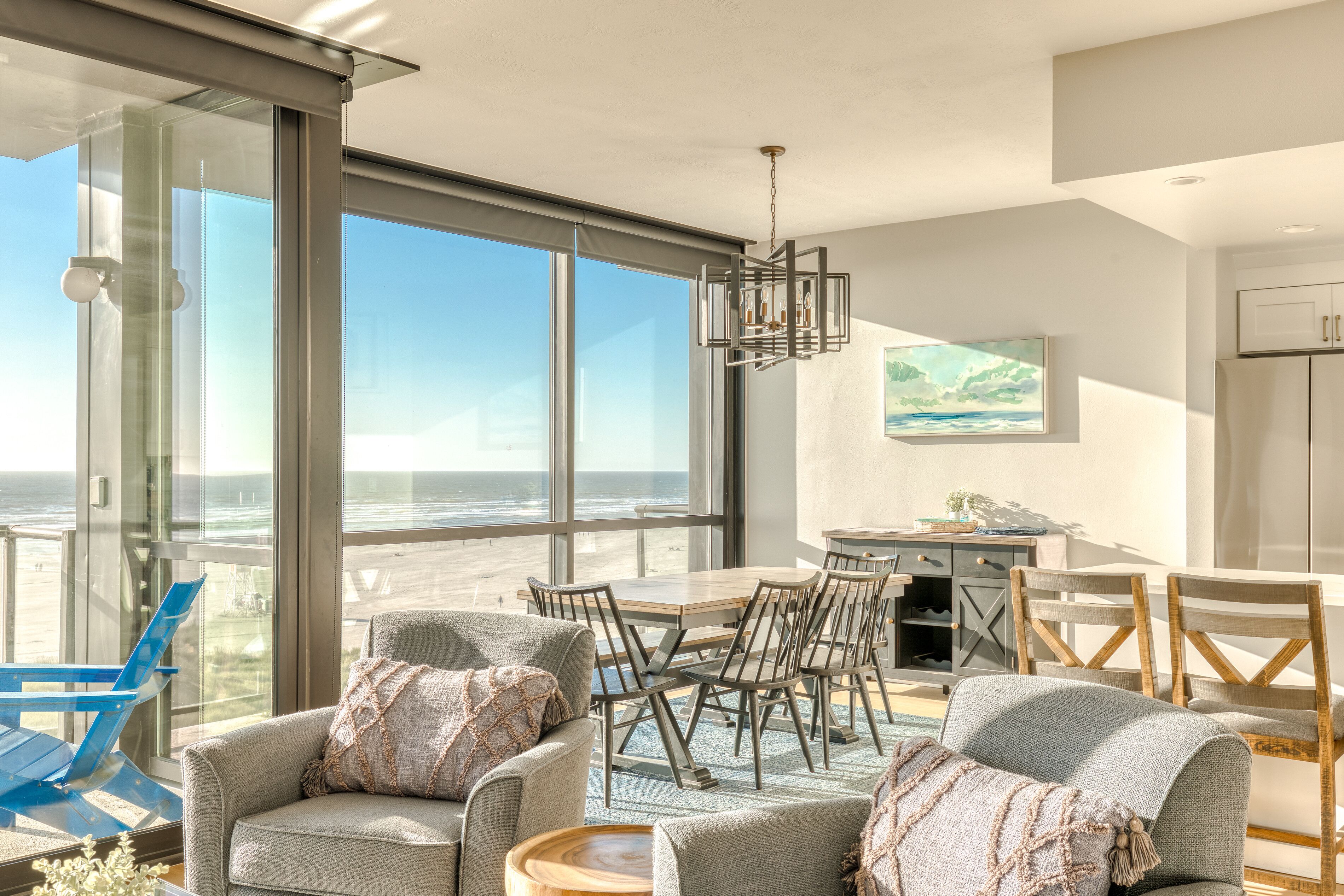 Un salon moderne doté d’une grande fenêtre donnant sur la plage, un coin repas avec une table et des chaises, et une cuisine équipée de placards et ornée d’un tableau.