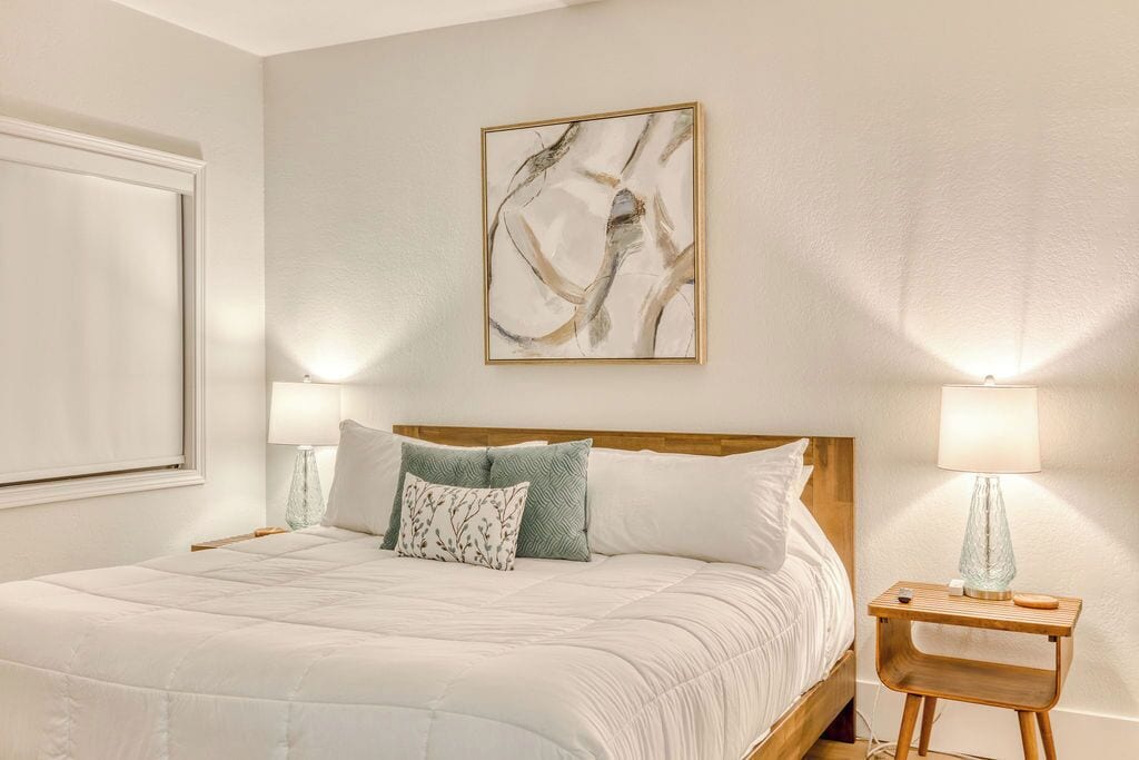 Ein ordentlich bezogenes Bett mit weißer Tagesdecke und mehreren Kissen, ein hölzernes Kopfende und ein Nachttisch mit Lampe.