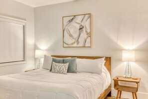 A neatly made bed with a white comforter and multiple pillows, a wooden headboard, and a bedside table with a lamp. - The Sand & Sea Condos - Oceanfront (Seaside)