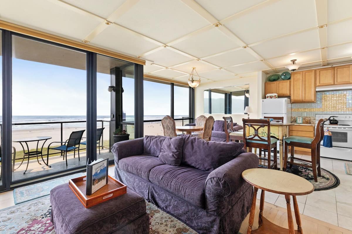 Un'ampia zona giorno con un grande divano viola, un tavolo da pranzo e sedie. È presente un angolo cottura con pensili in legno e un fornello. Ampie finestre offrono vista sulla spiaggia.