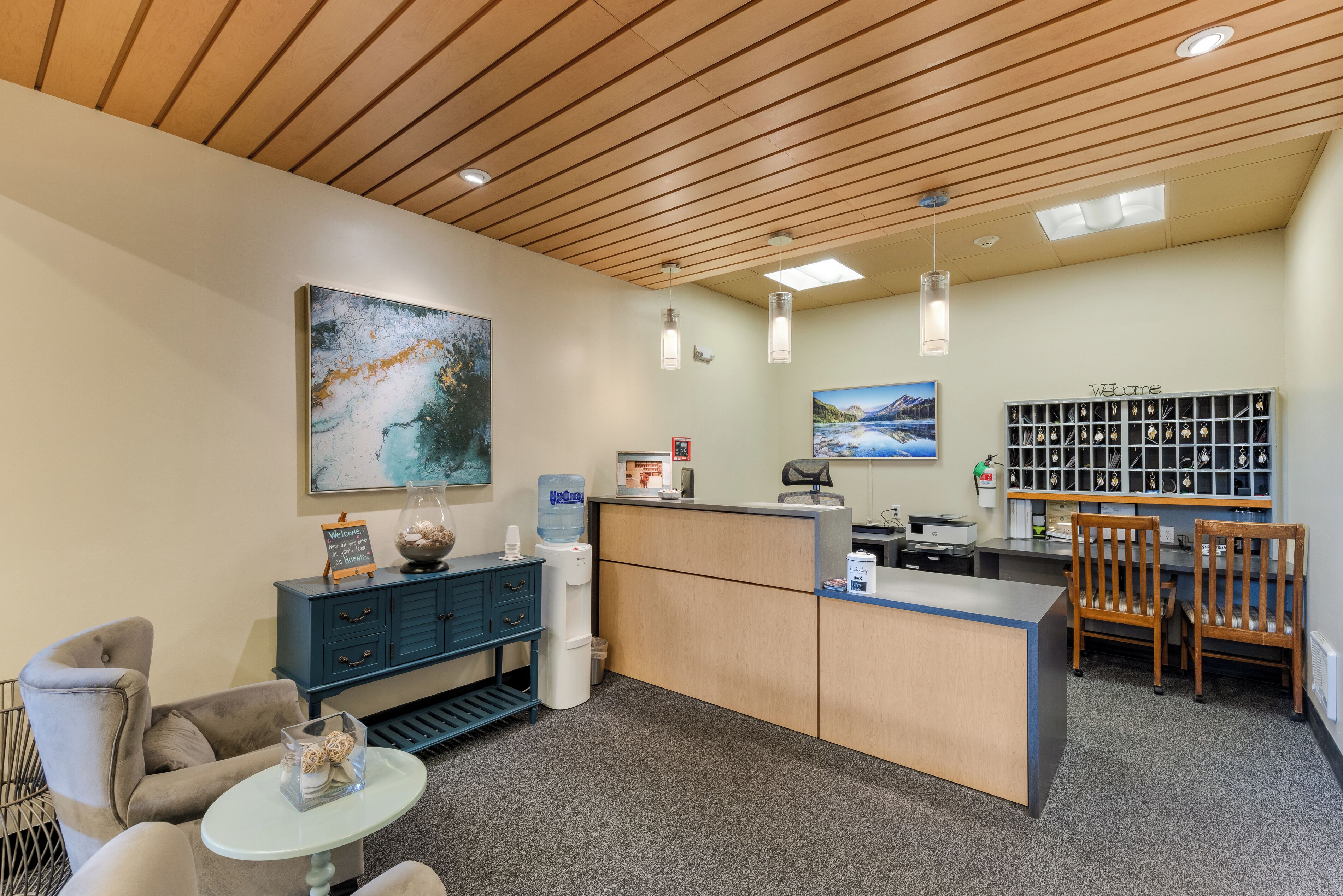 A reception area with a wooden ceiling, a reception desk, a water dispenser, a blue cabinet, a coffee machine, and a wall-mounted photo display.
