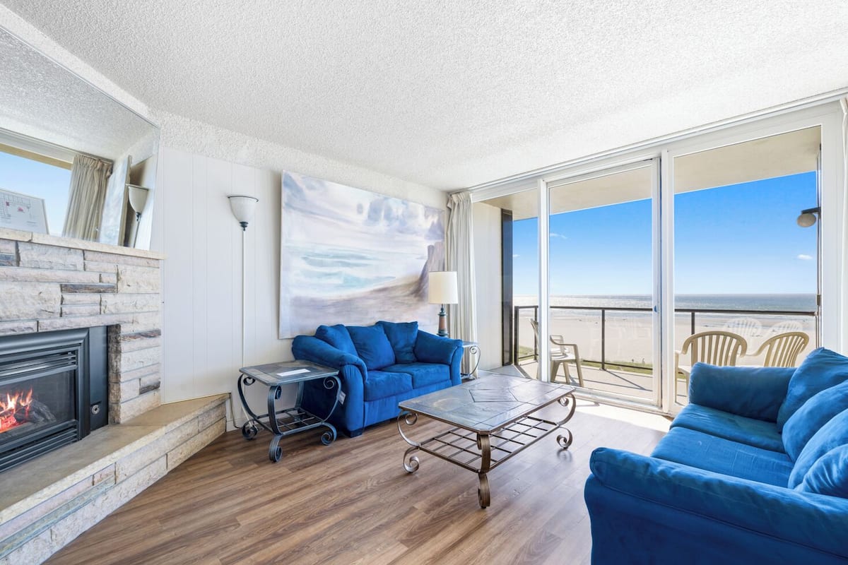 Un soggiorno moderno con camino, un divano blu, un tavolino da caffè e un balcone con vista sulla spiaggia.