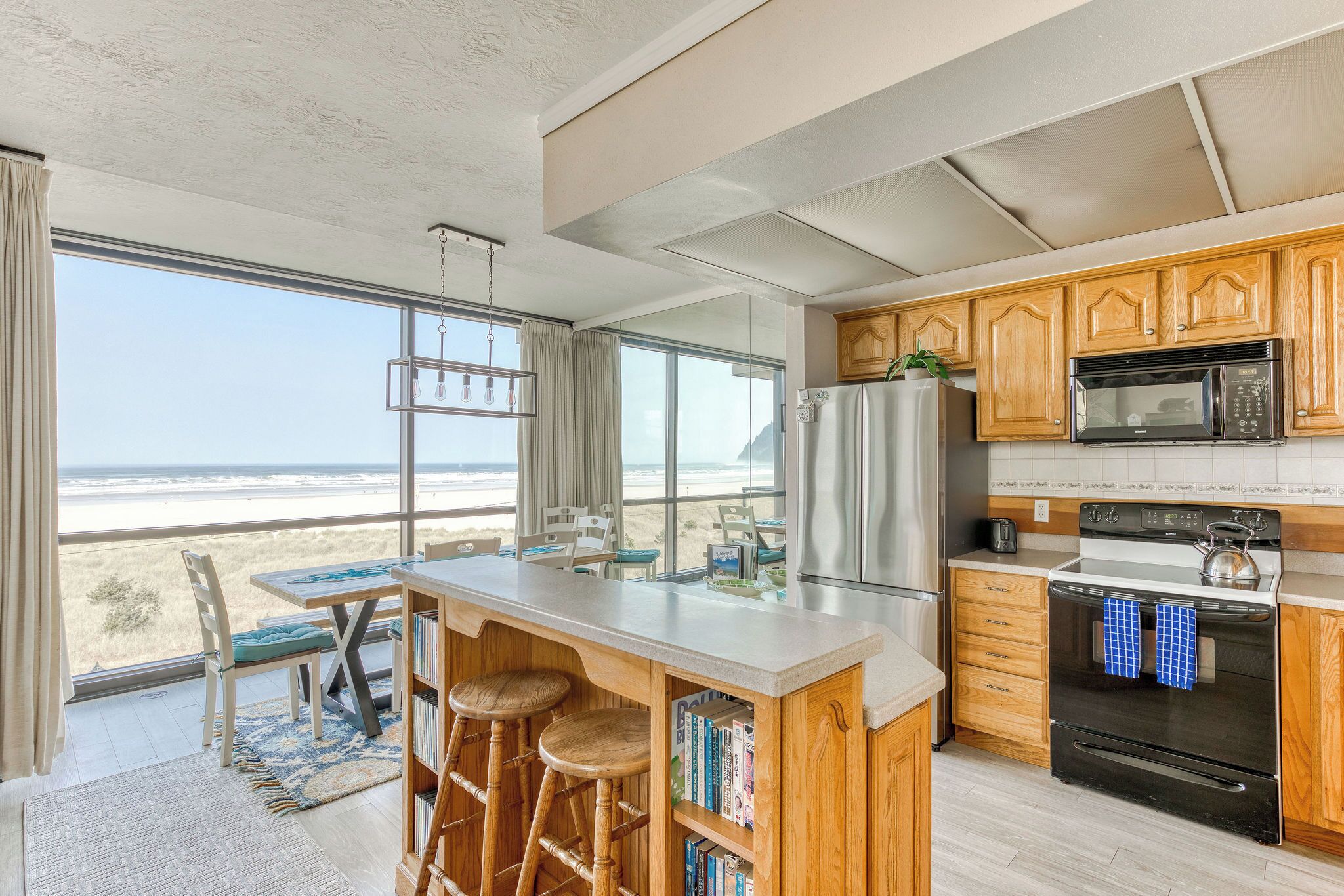 A kitchen with wooden cabinets, a stainless steel refrigerator, and a black oven. There is a large window with a view of the beach.
