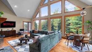 A spacious living room with a large sectional sofa, a dining table with chairs, and a flat-screen TV mounted on the wall. - Après Chalet|Ski|Luxury|Sauna|HotTub|Firepit|Disco (Windham)