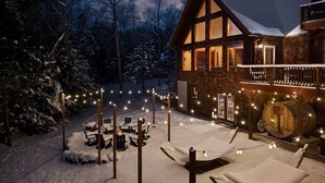 A cozy cabin with a snow-covered deck, outdoor seating, and a warm interior light. - Après Chalet|Ski|Luxury|Sauna|HotTub|Firepit|Disco (Windham)