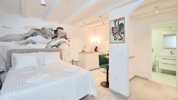 A hotel room with a bed, a small desk, a chair, a bathroom with a mirror, and artwork on the walls. - Karma Suites by Dominium Studio ground floor (Dubrovnik)