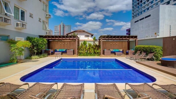 Una piscina con sillas de descanso y una zona de estar techada, rodeada de vegetación y edificios modernos.