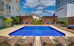 A swimming pool with lounge chairs and a covered seating area, surrounded by greenery and modern buildings. - GoldMen Business Joinville (Joinville)