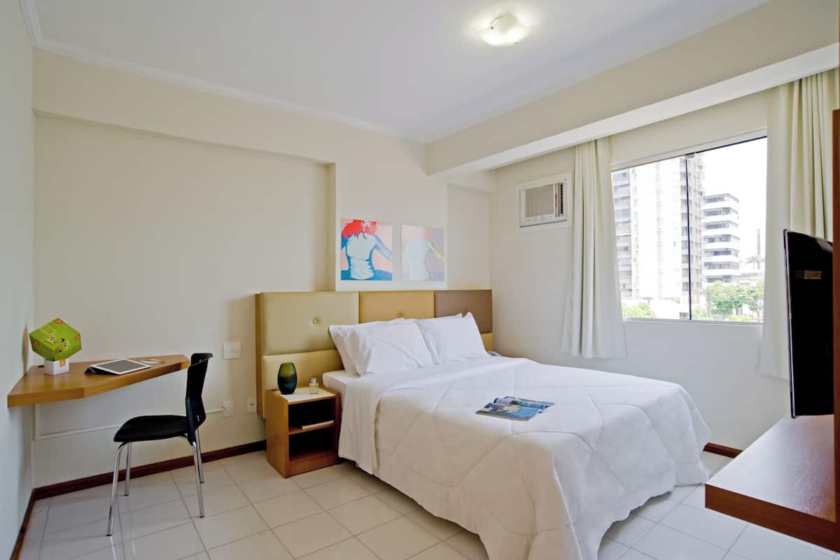 Quarto de hotel com cama, escrivaninha, cadeira, mesa de cabeceira e janela com vista para prédios.