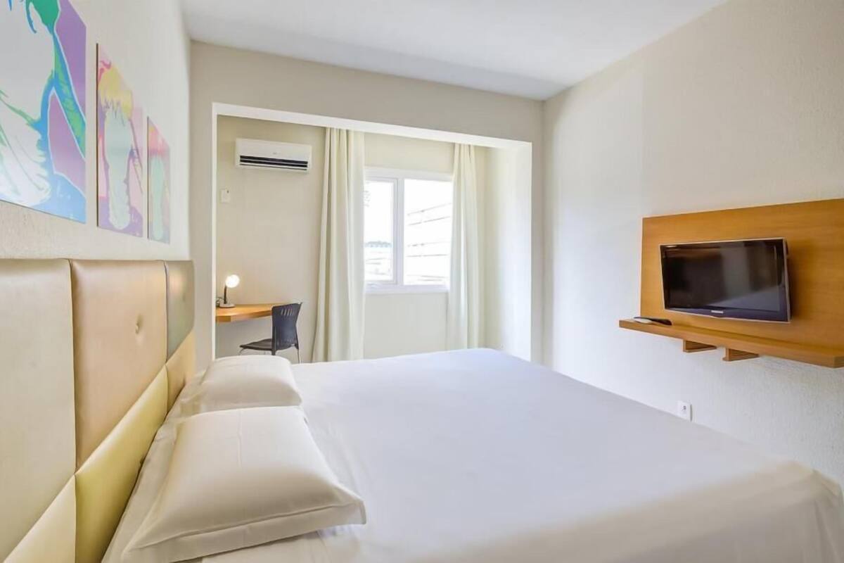 Habitación de hotel con cama, televisor montado en la pared, escritorio con silla y ventana con cortinas.