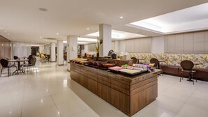 Un moderno hall de hotel con una zona de bufé central, un área de descanso con mesas y sillas, y un amplio ventanal con persianas.