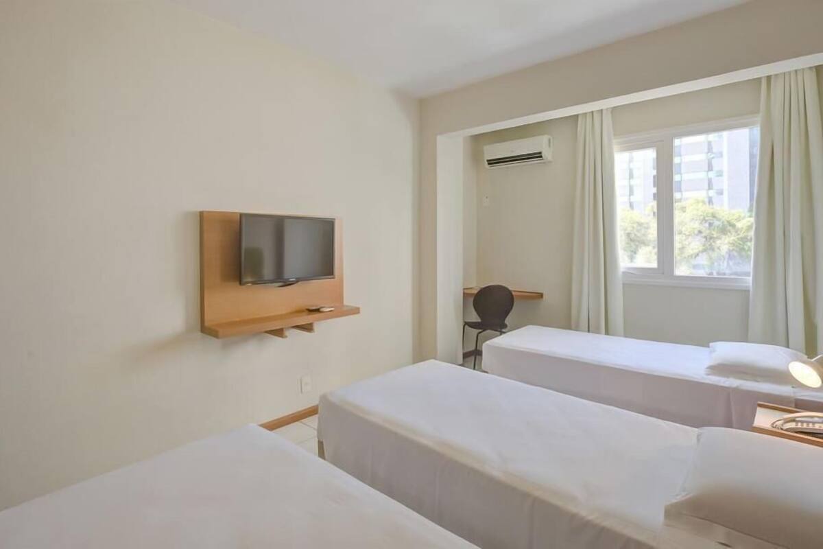 Una habitación de hotel con dos camas individuales, un televisor montado en un estante de madera, un aire acondicionado y una ventana con cortinas.