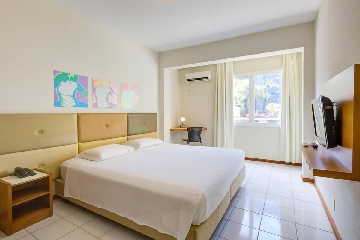 Habitación de hotel con una cama grande, una mesita de noche con lámpara, televisión y una obra de arte en la pared.
