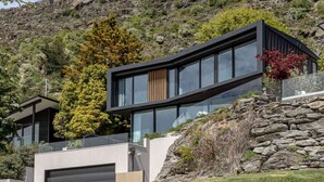 A modern house with a curved glass facade, a stone retaining wall, and a garage. - Willow Waters | Lakeside luxury with all day sunshine & fantastic outdoor living (Kawarau Falls)