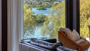 A modern living room with a large window offering a scenic view of a lake, mountains, and a town. - Willow Waters | Lakeside luxury with all day sunshine & fantastic outdoor living (Kawarau Falls)